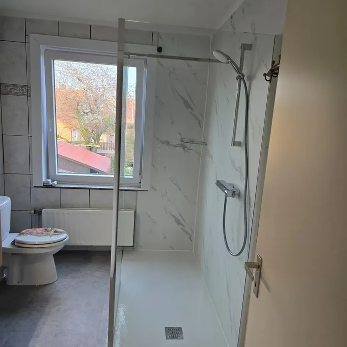 Inloopdouche met glazen wand en witte marmerlook wandpanelen, staand toilet en raam op de achtergrond