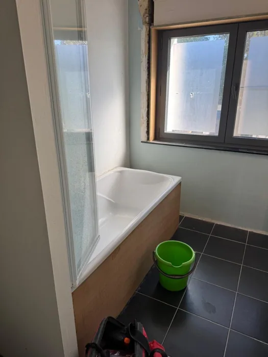 Oude badkamer met ligbad en badwand onder raam, zwarte vloertegels